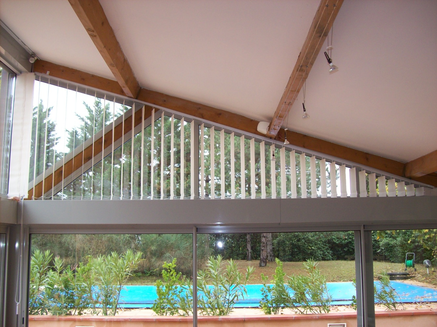 Véranda avec fenêtre inclinée équipée de stores verticaux blancs, vue sur jardin et piscine bleue.