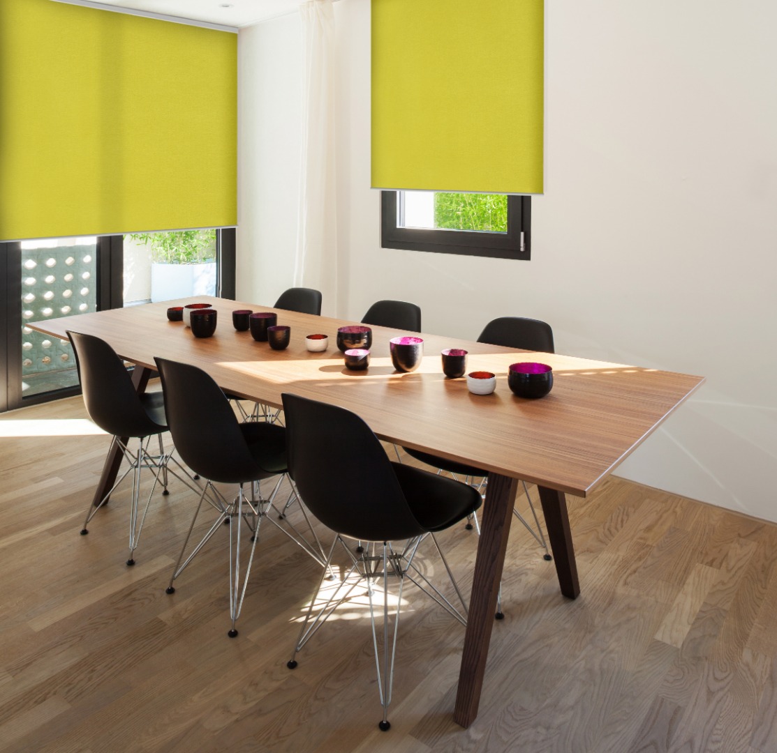 Salle à manger lumineuse avec table en bois, chaises noires et stores enrouleurs jaunes vifs aux grandes fenêtres.
