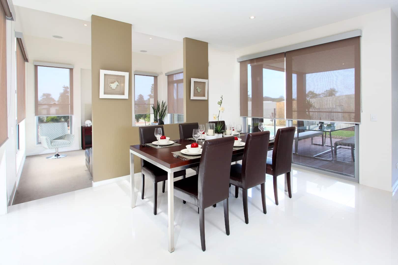 Salle à manger moderne avec table en bois, chaises en cuir marron, stores enrouleurs marron et sol carrelé blanc brillant.