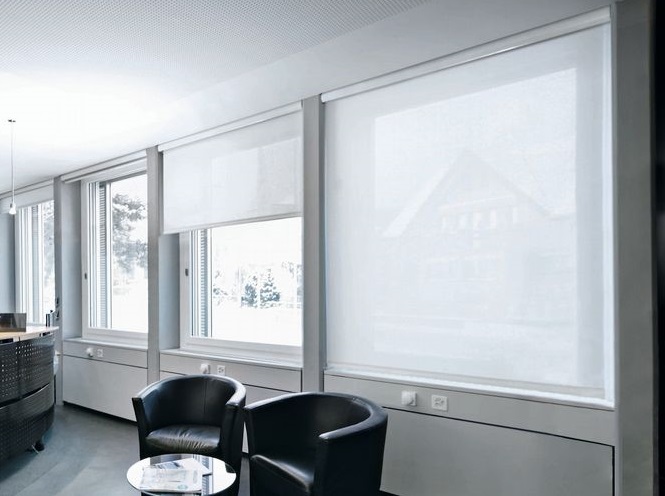 Intérieur de bureau lumineux avec larges fenêtres équipées de stores à enrouleur blancs, fauteuils noirs, table basse.