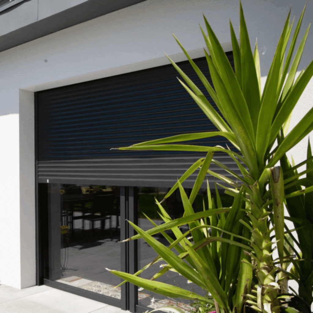 Gros plan sur un volet roulant noir partiellement fermé protégeant une baie vitrée d'une maison moderne, encadré par un grand yucca vert.