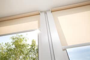 Store enrouleur beige sur fenêtre, lumière du soleil à travers arbre, ciel bleu. Vue de près sur les stores installés.