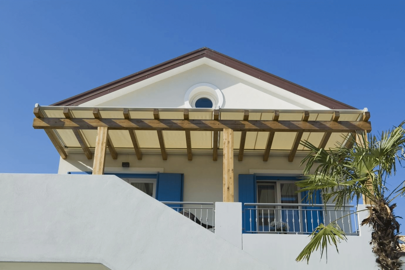 Façade de maison blanche méditerranéenne sous ciel bleu, toit marron, oculus, pergola en bois clair avec store et volets bleus.