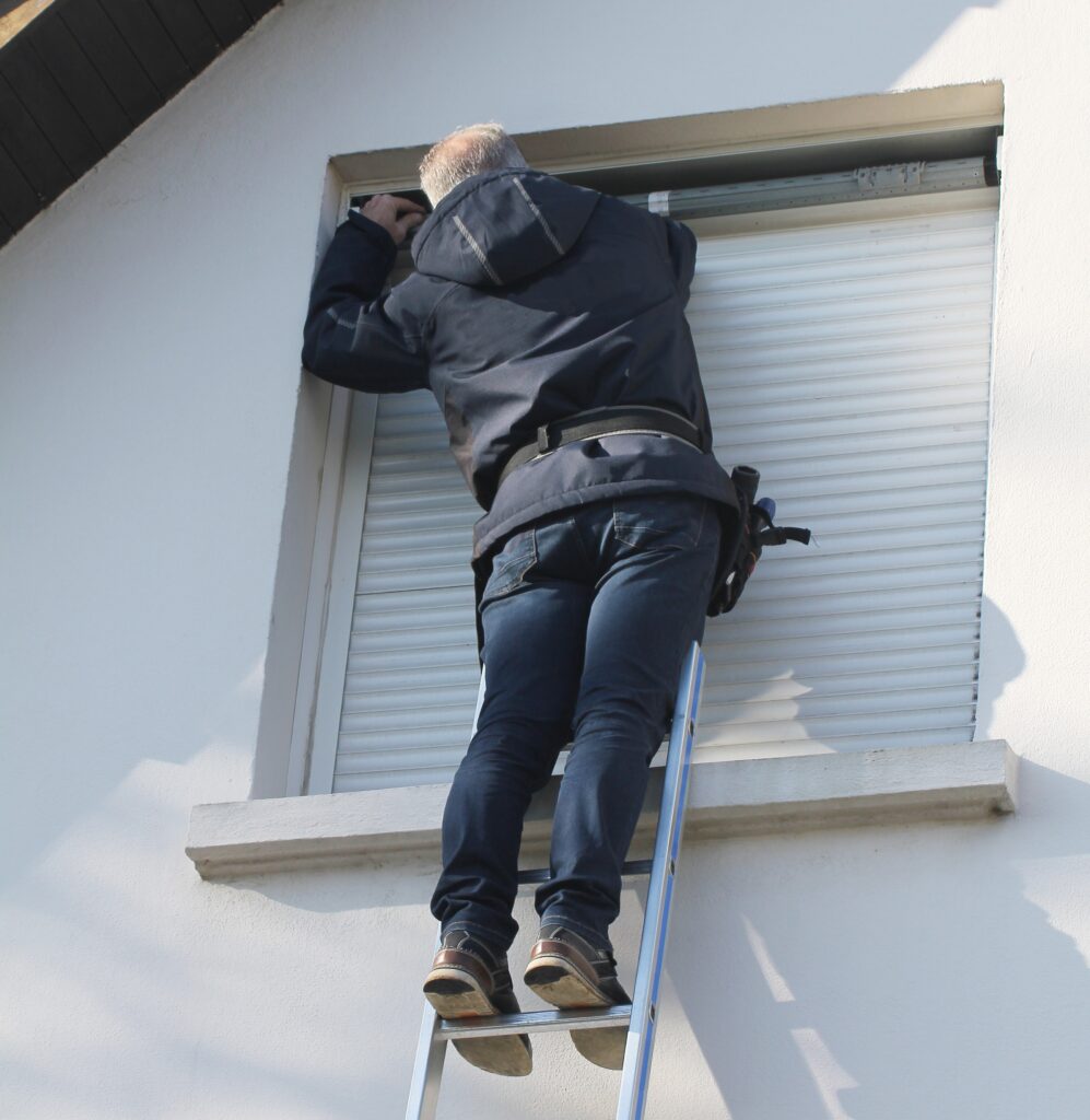 Réparation de volet roulant : un expert sur échelle Homme sur une échelle réparant le mécanisme d'un volet roulant sur une fenêtre. Mur extérieur blanc ensoleillé.