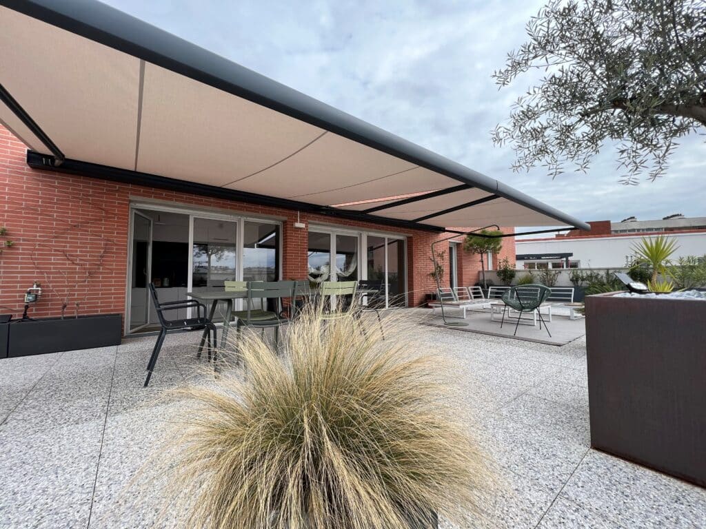 Terrasse moderne design : pergola rétractable, salon jardin, plantes Grande terrasse moderne avec pergola rétractable beige, salon de jardin vert et gris, et plantes variées sur sol clair, façade en briques.