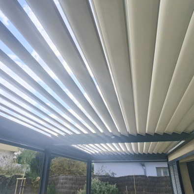 Toit de pergola bioclimatique à lames orientables blanches. L'ouverture partielle révèle le ciel bleu au-dessus d'une terrasse.