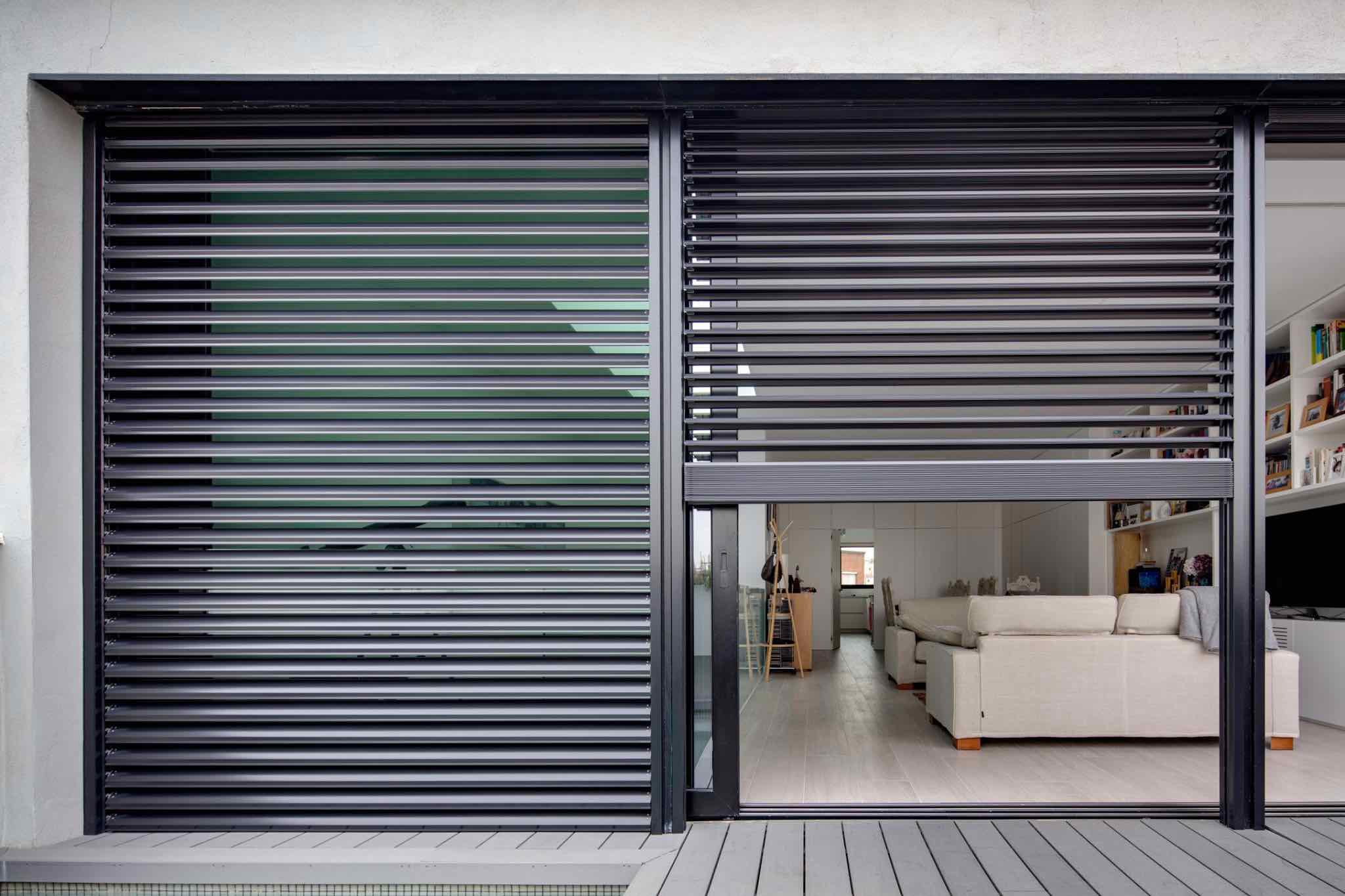 Brise-soleil aluminium noir sur porte-fenêtre coulissante. Le panneau droit est ouvert, révélant l'intérieur lumineux d'un salon.