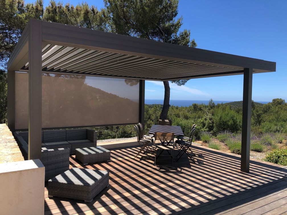 Pergola bioclimatique moderne : aménagement de terrasse avec vue mer. Pergola bioclimatique grise sur terrasse en bois. Salon de jardin, store latéral et vue mer imprenable sur la nature sous un ciel bleu.