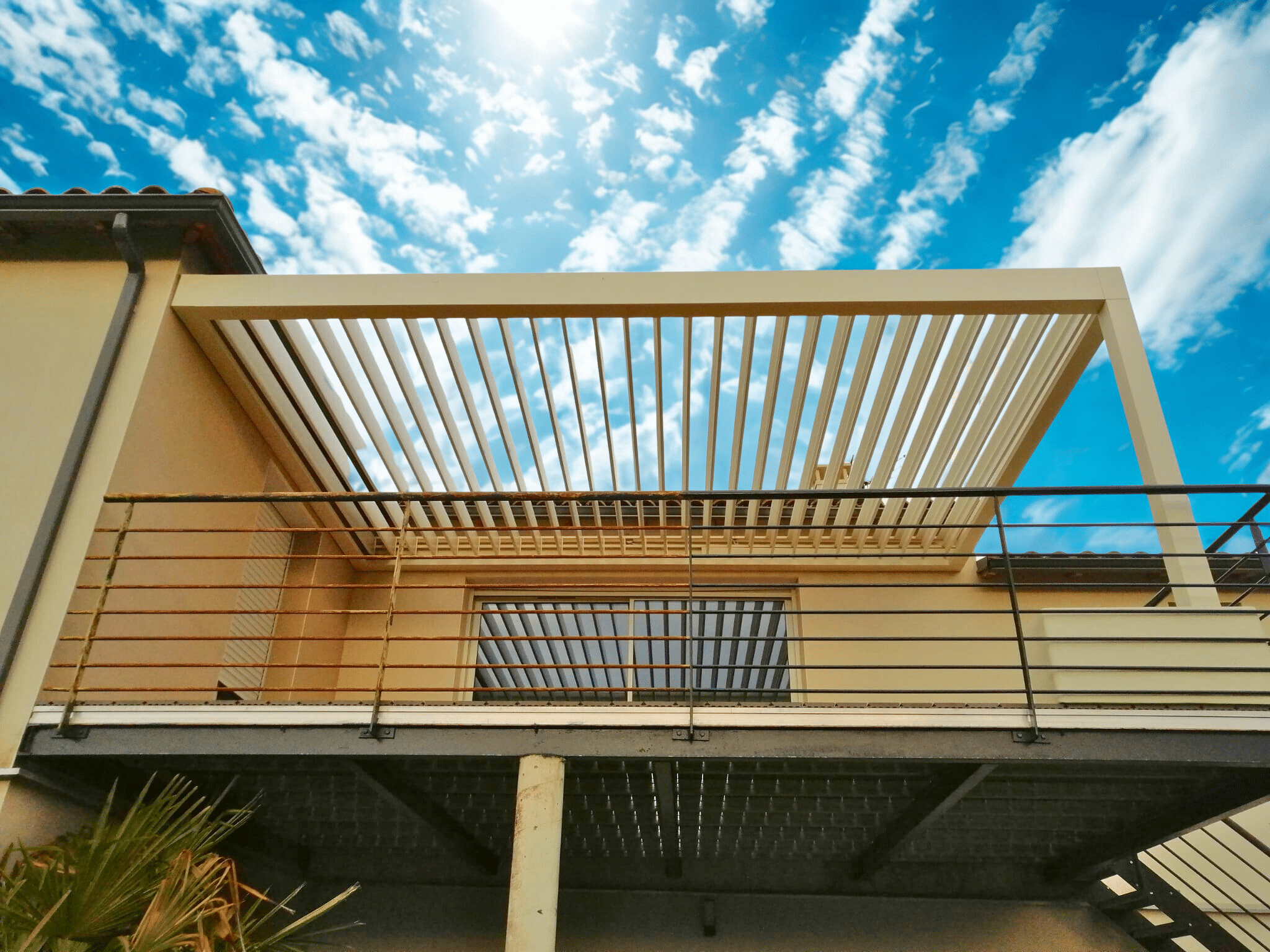 Pergola bioclimatique beige moderne fixée à la façade et sur pied, protégeant une terrasse. Lames orientables et garde-corps en métal.
