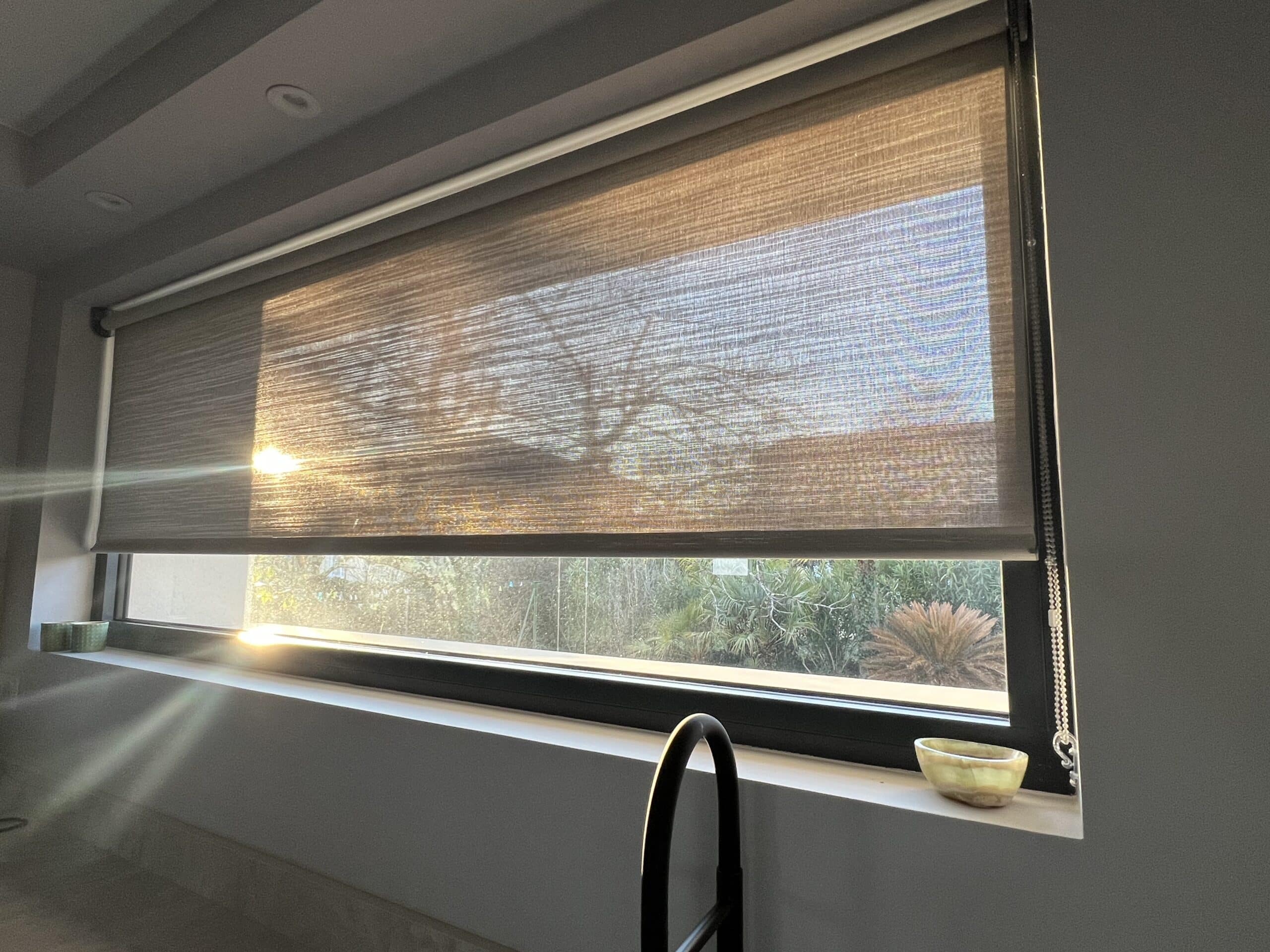 Store enrouleur filtrant la lumière du soleil couchant sur une fenêtre d'intérieur moderne. Arbres visibles en transparence.