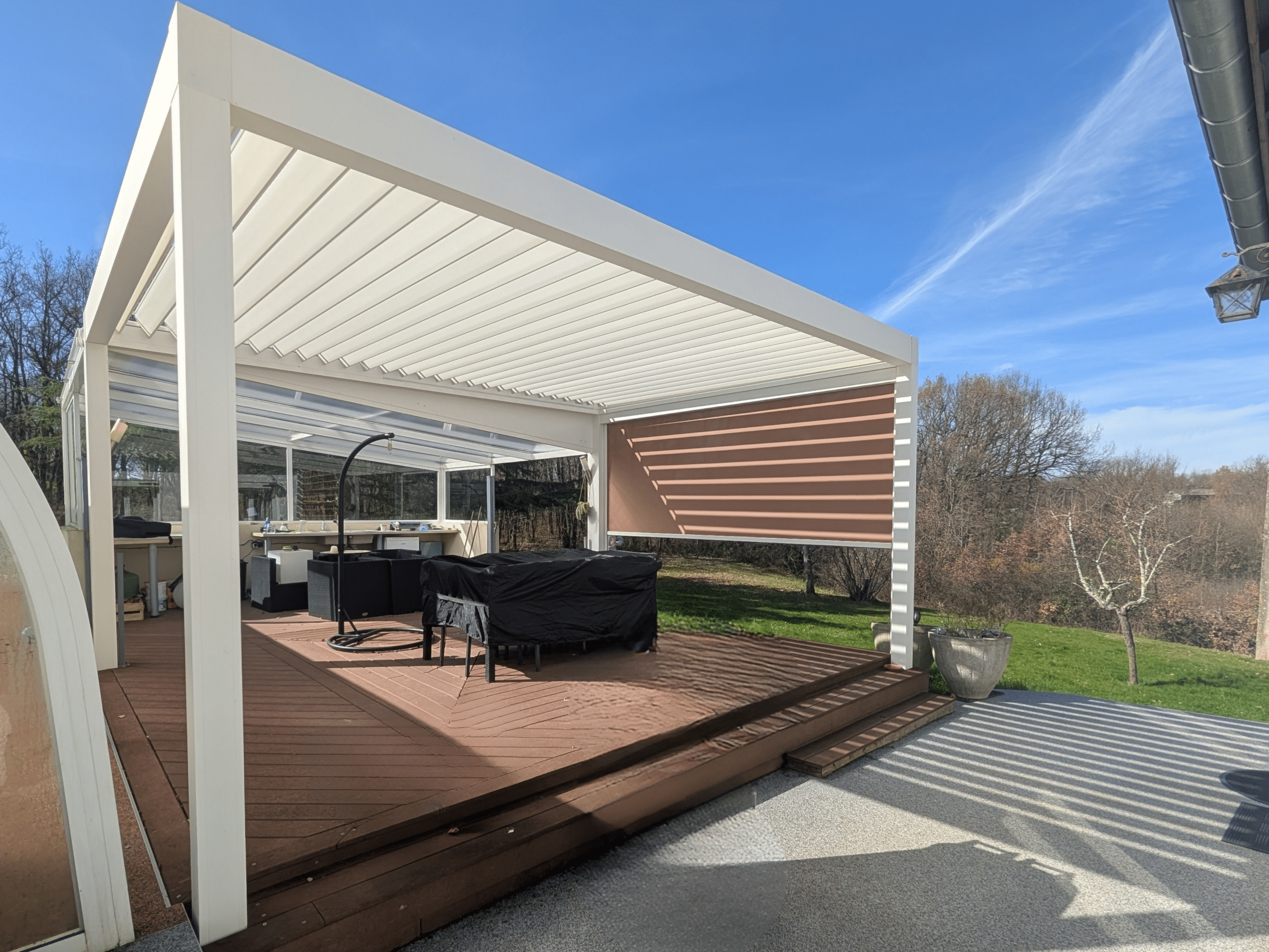 Pergola bioclimatique blanche sur terrasse en bois avec store latéral et mobilier de jardin sous un ciel bleu.