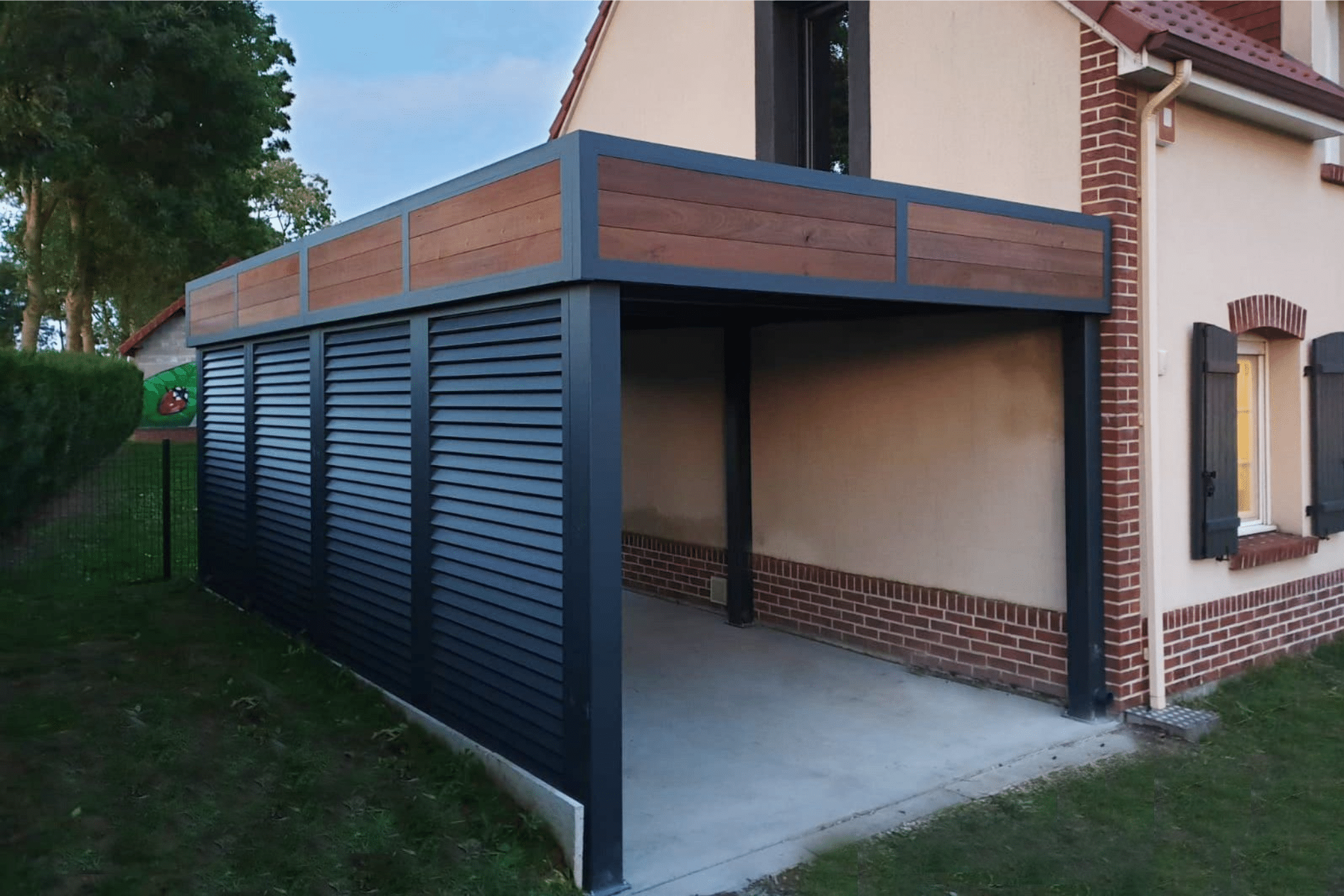 Carport moderne gris anthracite avec toit en bois et parois à persiennes, adossé à une maison avec briques.