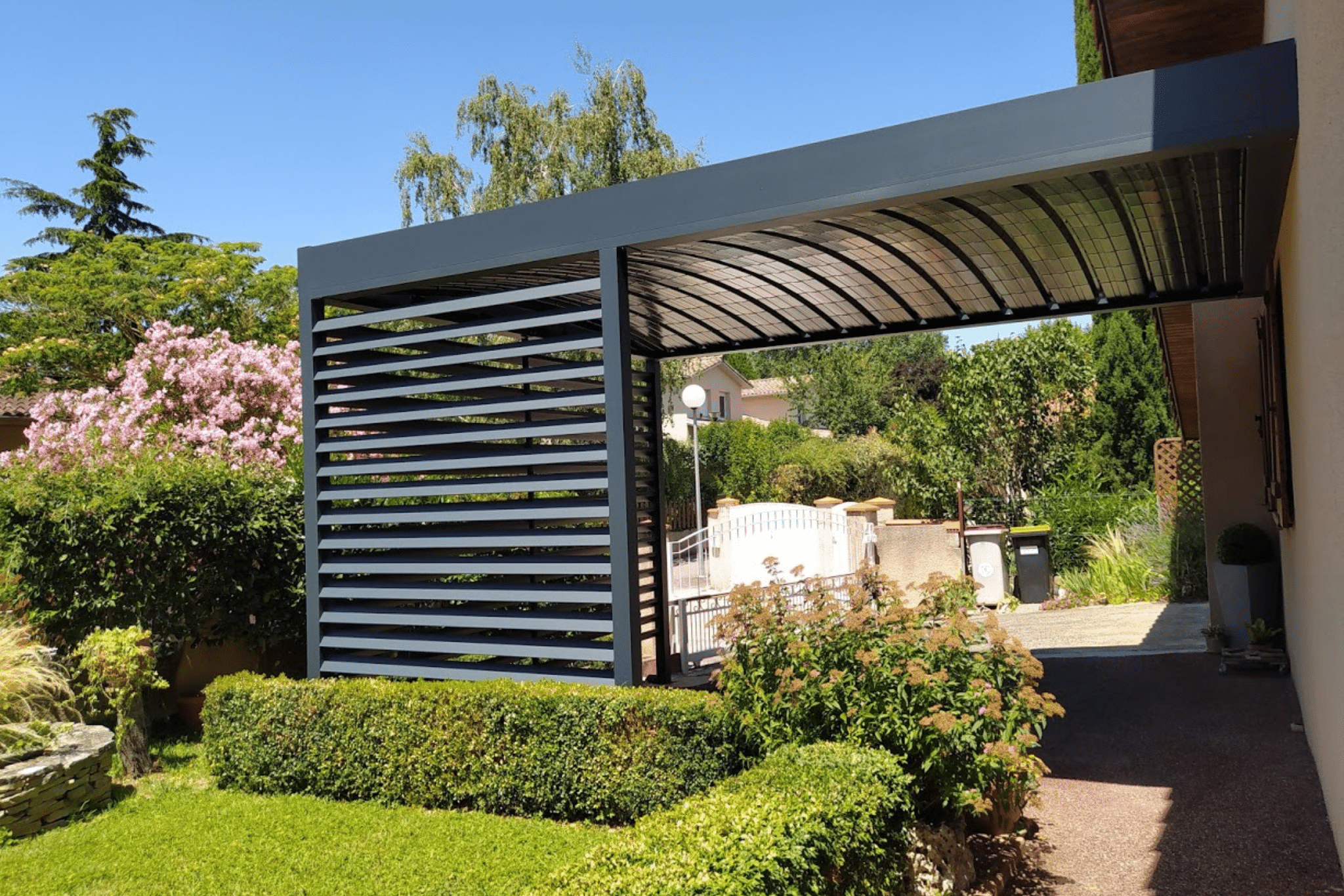 Carport moderne en aluminium gris anthracite avec toit incurvé et brise-vue à lames dans un jardin fleuri.