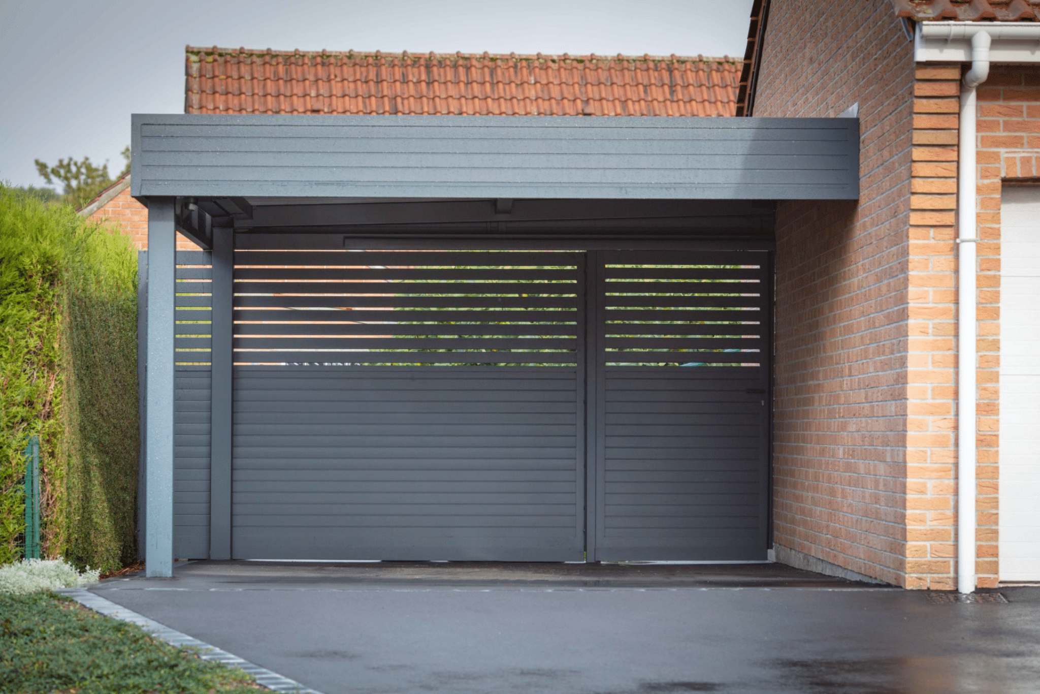 Carport moderne gris anthracite avec portail à lattes devant une maison en briques et allée bitumée.