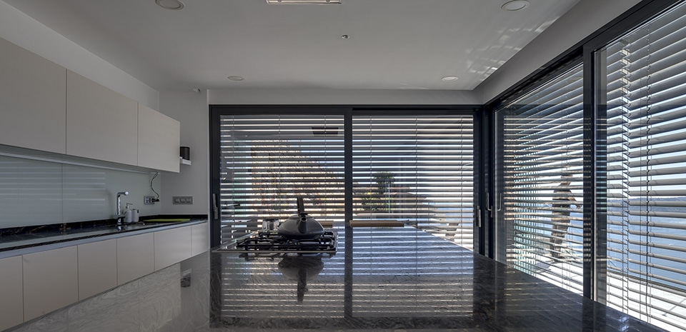 Cuisine moderne avec îlot en marbre noir, gazinière, et grandes baies vitrées avec stores offrant une vue sur la mer.