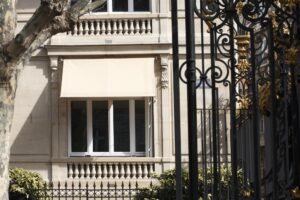 Façade classique avec store banne beige, balustrade en pierre et grille en fer forgé noire et dorée.