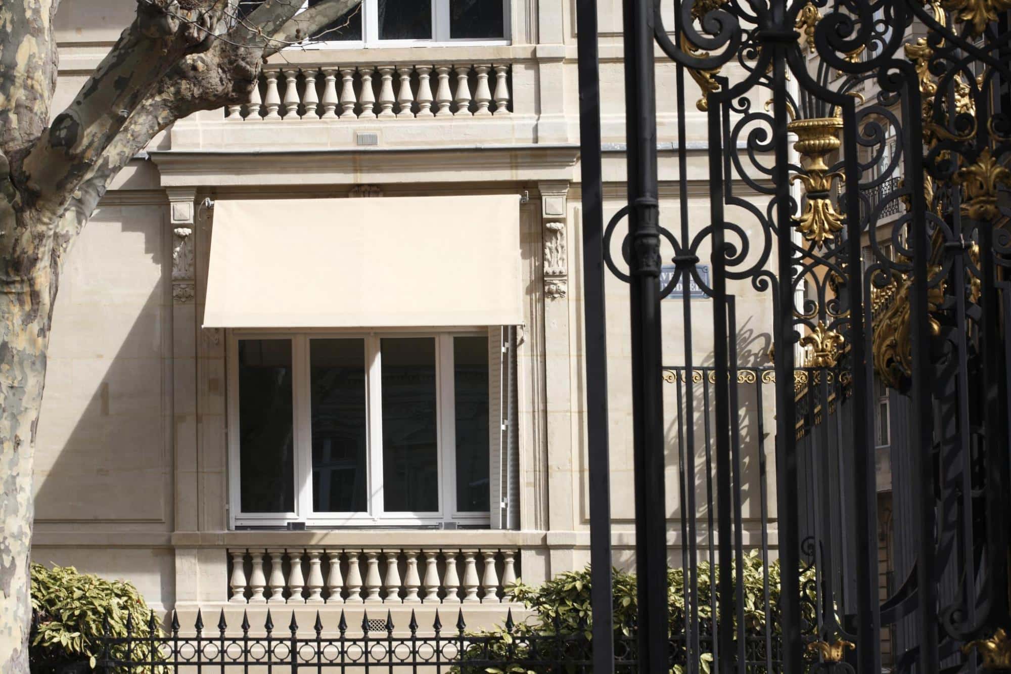 Façade classique avec store banne beige, balustrade en pierre et grille en fer forgé noire et dorée.