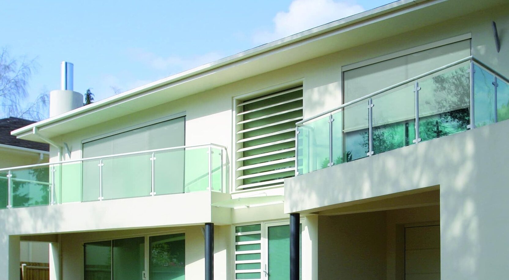 Maison moderne blanche à deux étages avec balcons en verre, allée pavée et jardin paysager sous un ciel bleu clair.