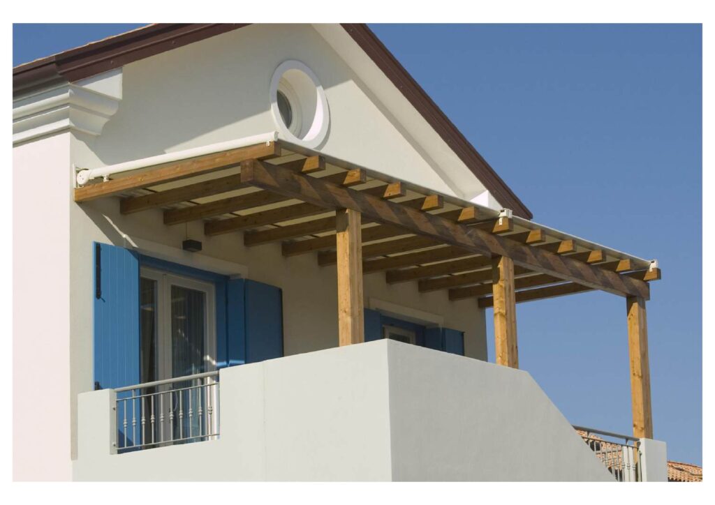 Pergola en bois et store rétractable pour balcon de maison Pergola en bois avec store rétractable blanc sur le balcon d'une maison aux volets bleus sous un ciel dégagé.