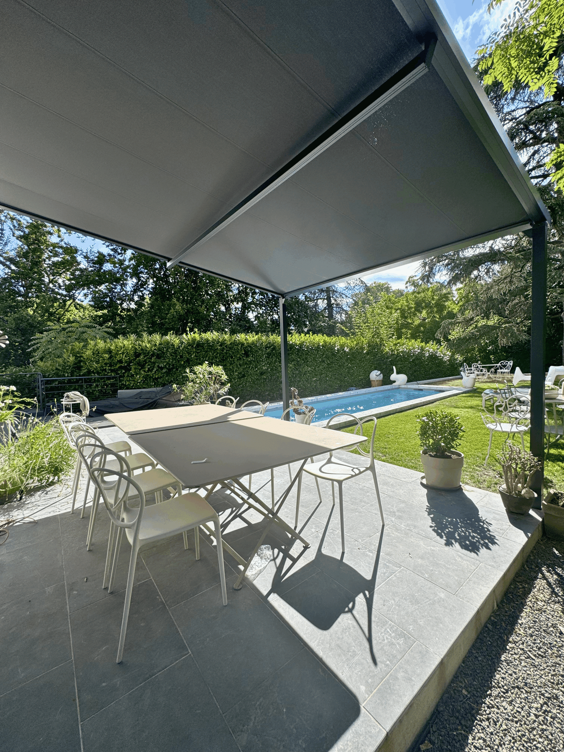 Terrasse élégante sous pergola grise avec mobilier de jardin blanc, piscine et jardin verdoyant en arrière-plan.