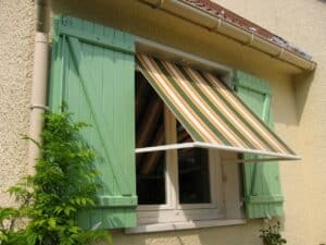 Fenêtre avec volets battants vert pastel et store banne rayé orange et vert sur une façade en crépi beige.