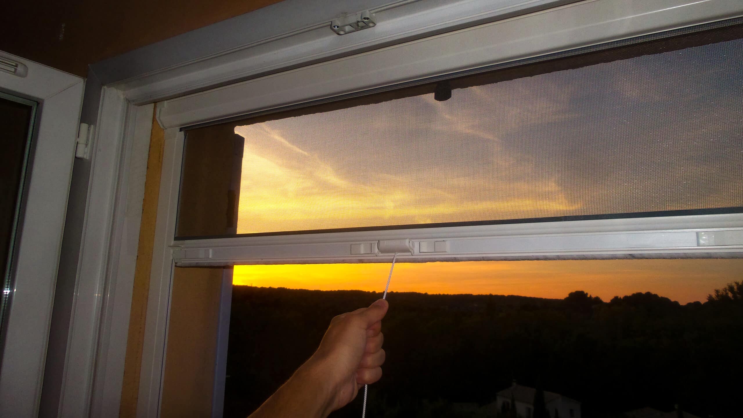 Main tirant une moustiquaire enroulable de fenêtre face à un magnifique coucher de soleil orangé sur la forêt.