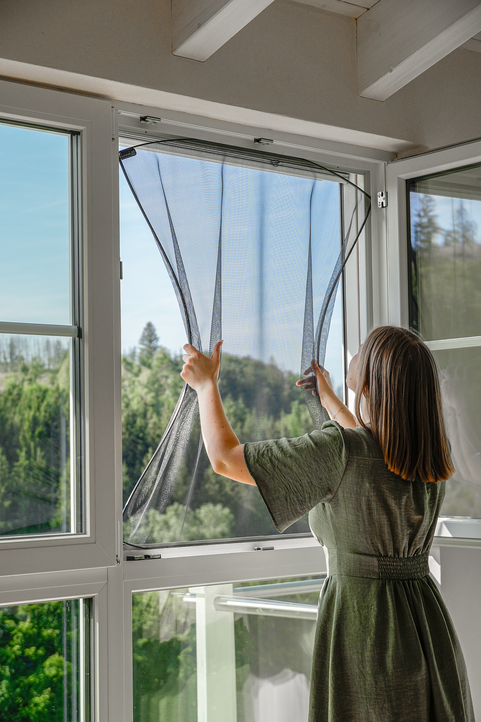 Femme installant une moustiquaire magnétique sur une fenêtre blanche avec vue sur une forêt verdoyante ensoleillée.