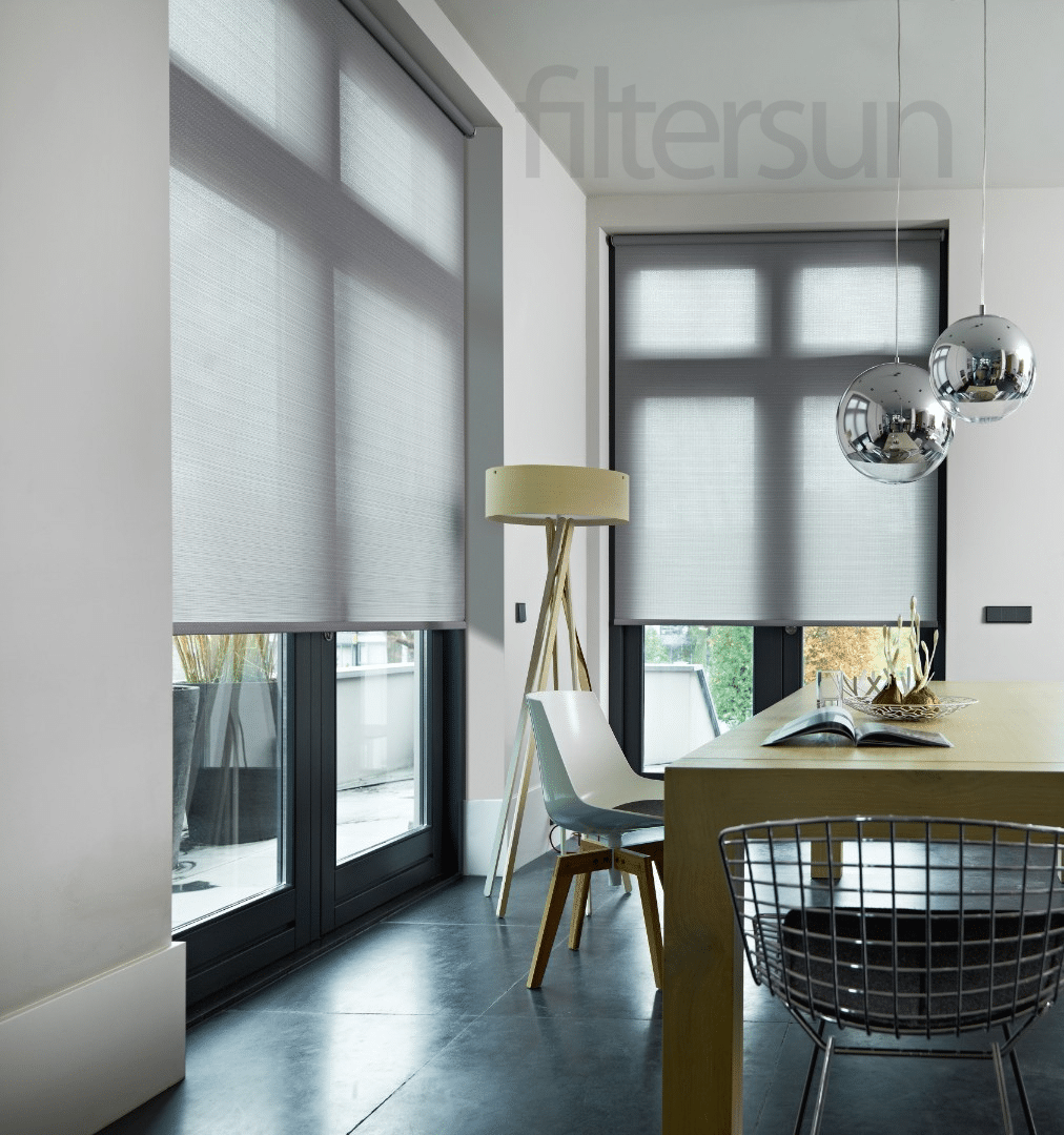 Store enrouleur gris Filtersun tamisant sur grandes fenêtres d'un salon moderne avec table en bois et chaises.
