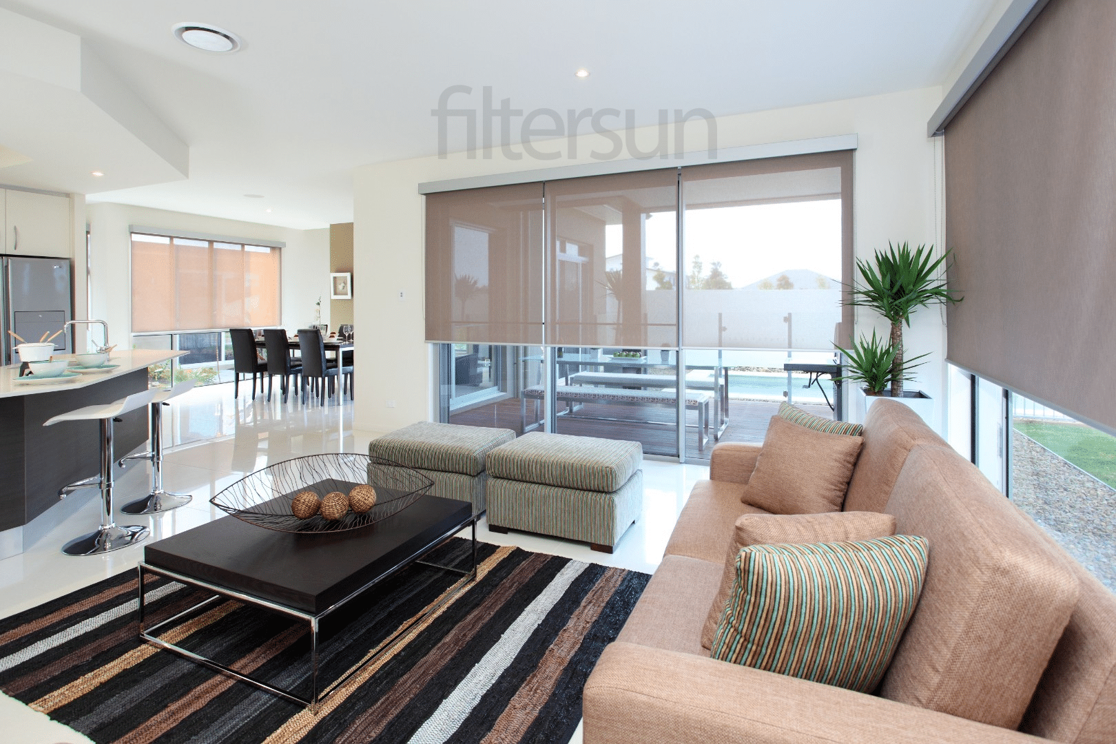 Salon moderne lumineux avec stores enrouleurs filtersun, canapé beige, tapis rayé et table basse design en bois sombre.