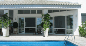 Terrasse avec piscine bleue, baies vitrées blanches avec moustiquaires et grandes jardinières de plantes grimpantes.